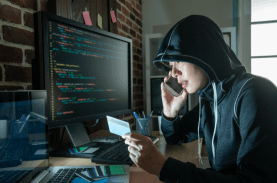 A hacker holding a credit card and speaking on the phone, with a computer displaying code in the background.